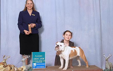 Judge Rachel Romano Kelly stands next to Wednesday Raymond and Johnson Ivyside Two Scoops Vanilly, holding a brown ribbon. There are gold and silver christmas decorations around the stand.